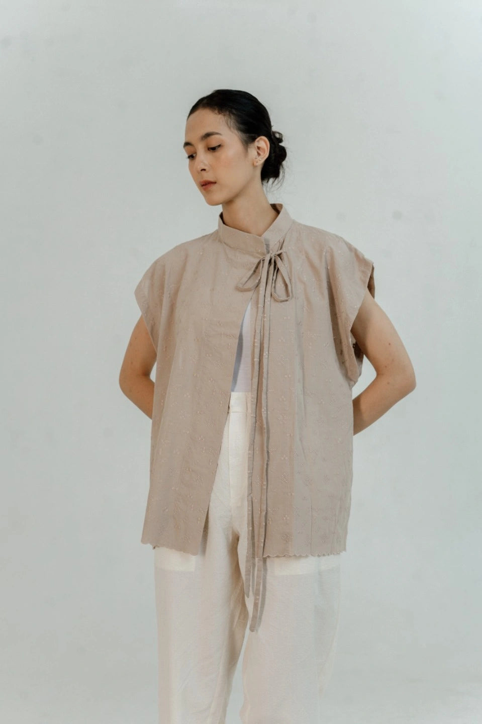 Woman wearing a beige sleeveless outer by Damakara with a tie neckline against a white background