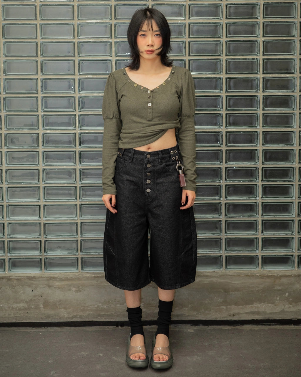 Person wearing a green long-sleeve top and black wide-leg jorts pants standing against a glass-block wall.