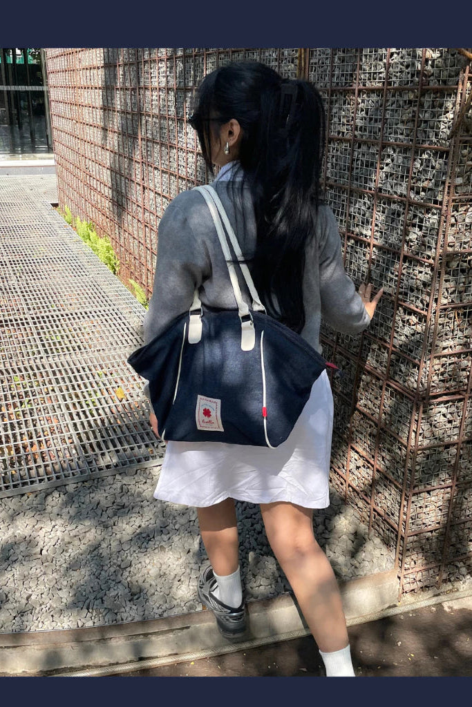 A woman viewed from behind wearing a gray long-sleeve top and white skirt, carrying a navy blue denim tote bag by Luella with white straps and a small red logo patch on a city walkway.