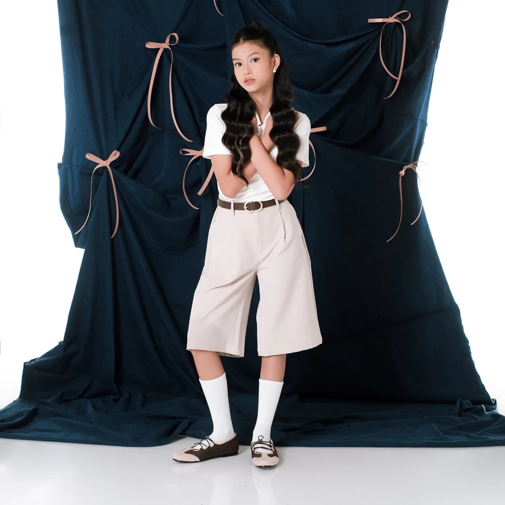 Woman in a white top, beige shorts, and flat shoes standing in front of a large dark blue curtain with pink ribbons.