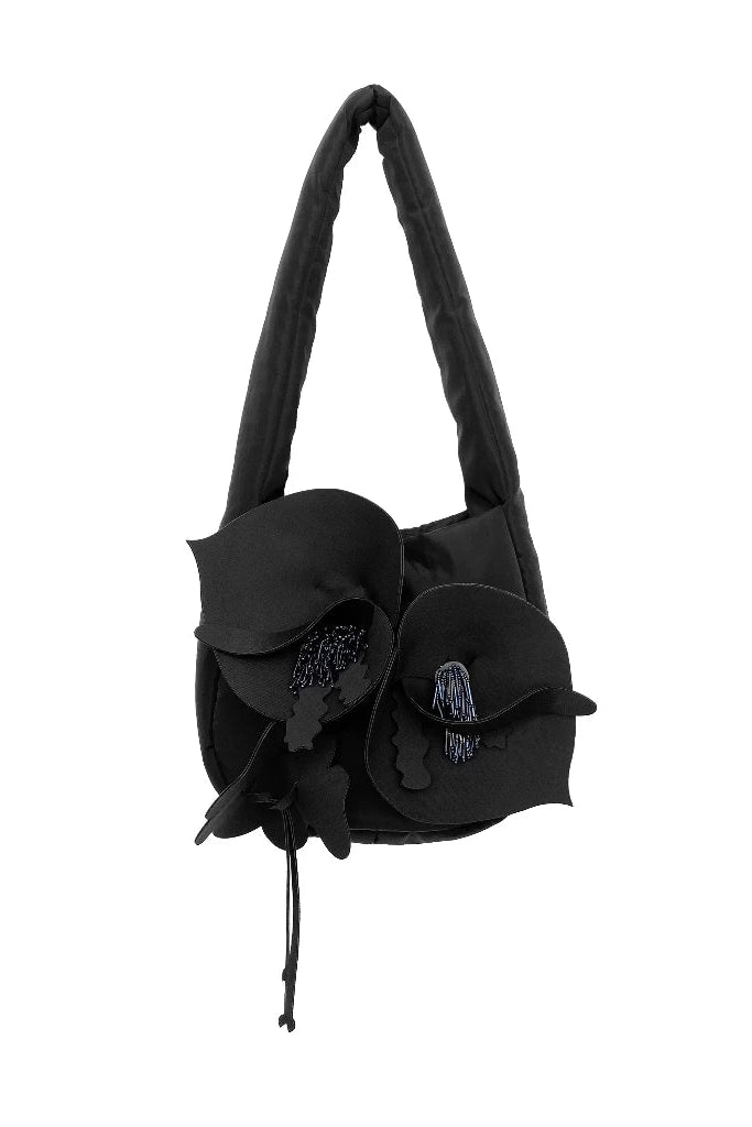 A black fabric shoulder bag by Hunting Fields with a long strap, featuring large sculptural flower-like fabric petals and a small hanging black tassel on a plain white background.