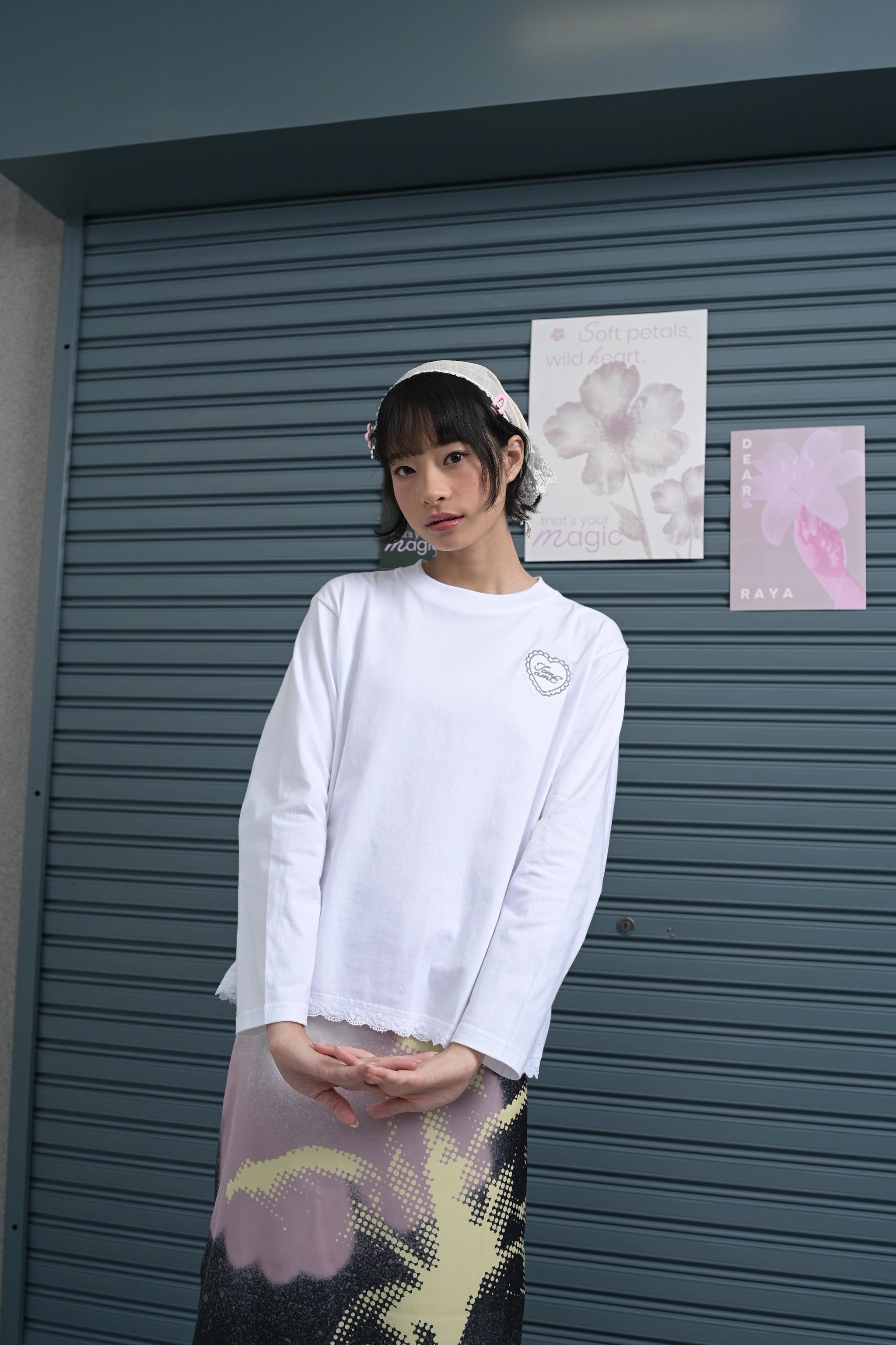 Person wearing a white long-sleeve t-shirt by Tome Ame with a heart design and lace details in front of a gray shutter.