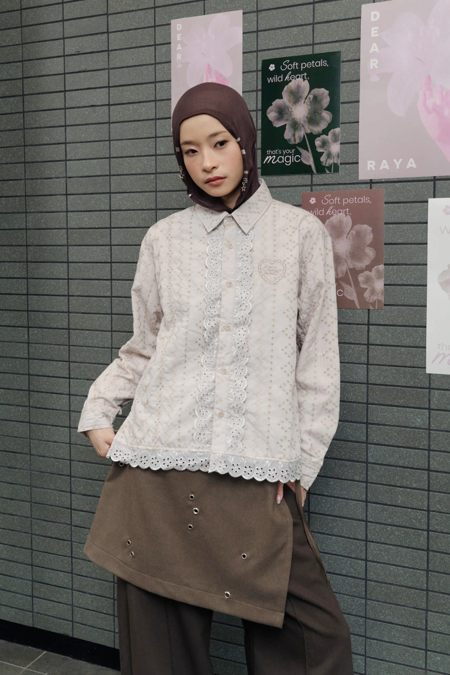Person wearing a light brown lace shirt by Tome Ame and brown pants against a tiled wall with posters.