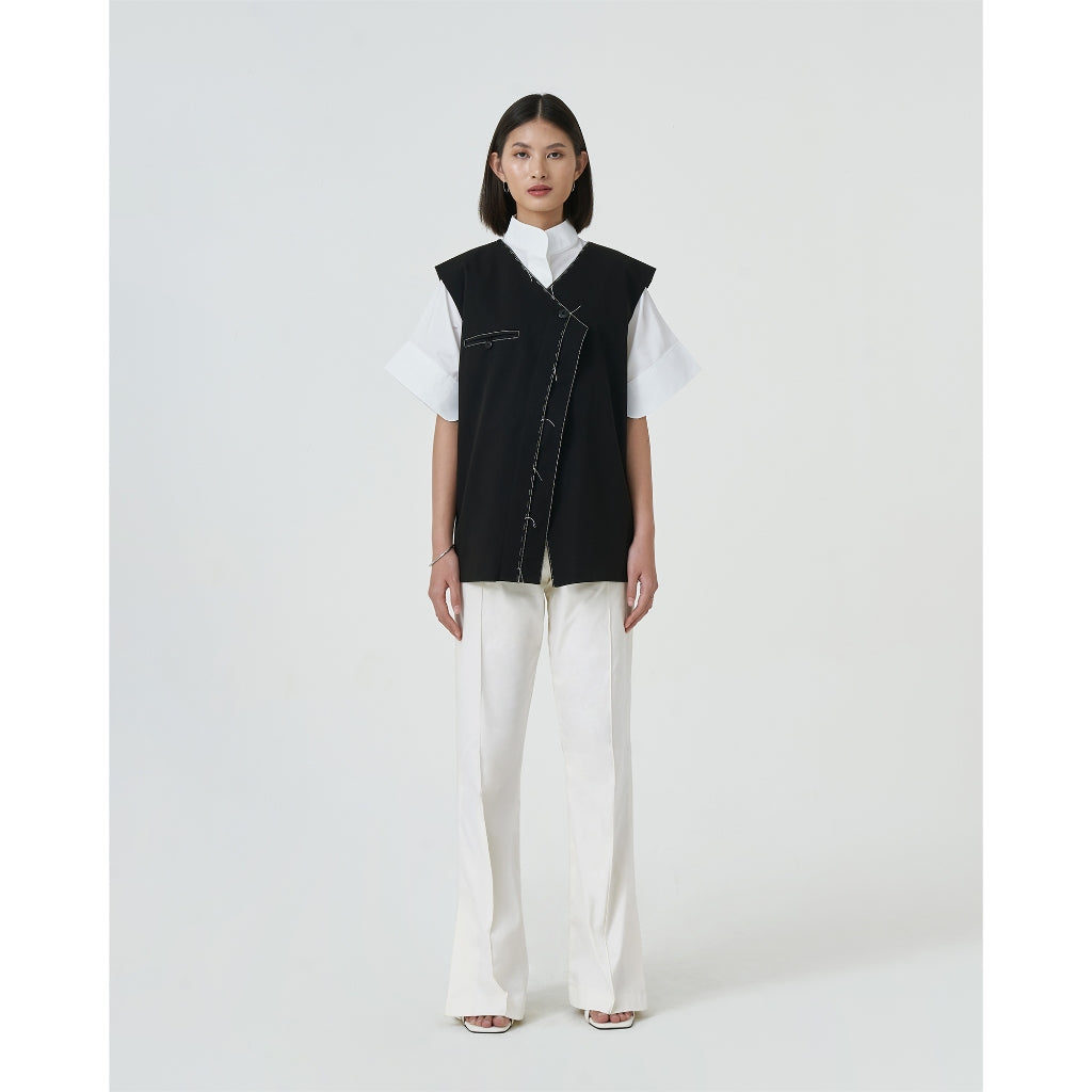 A woman wearing a black collarless vest with contrast handstitches details and white trousers. The vest has a hidden button on the inside.