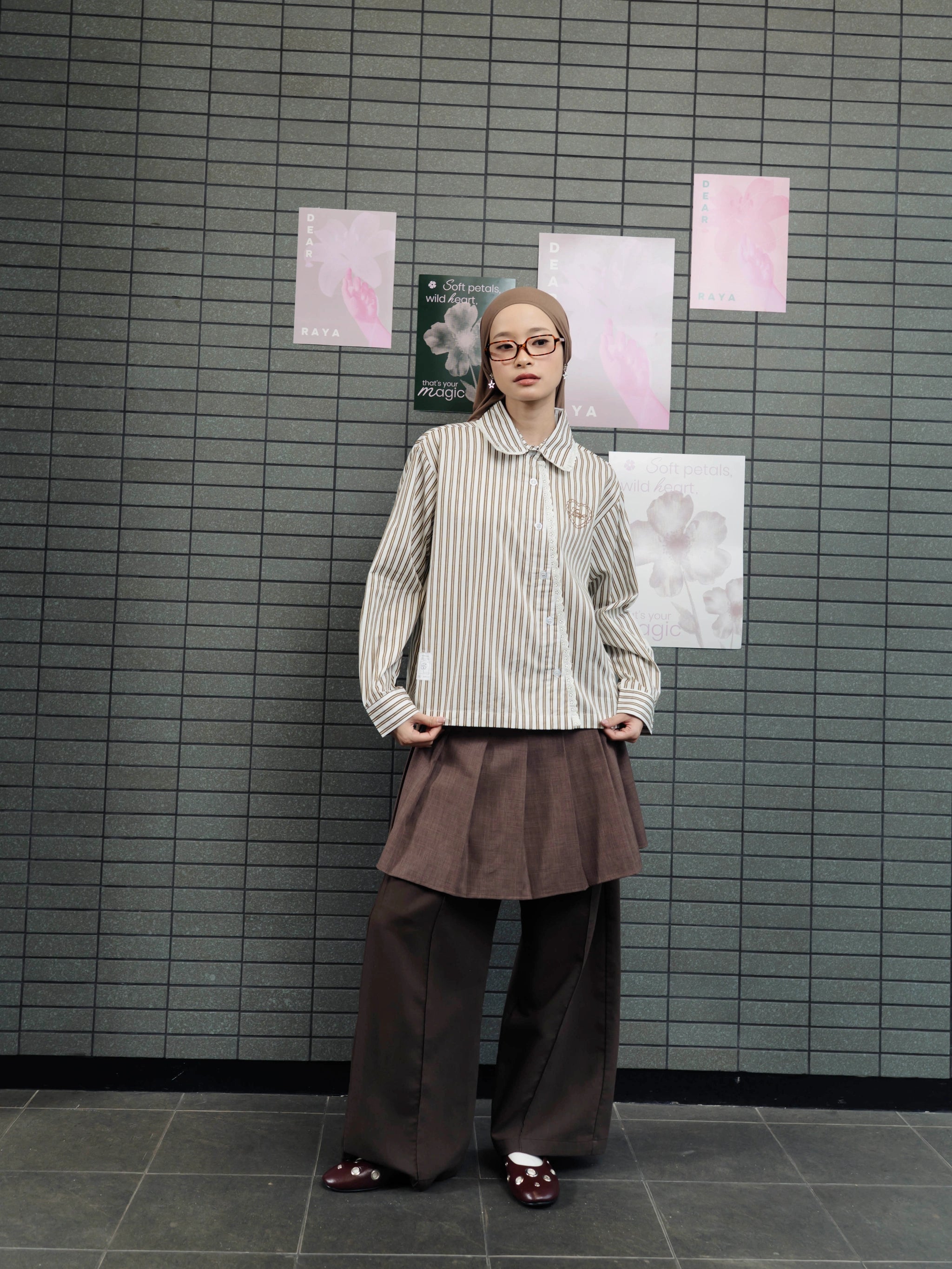Person wearing a brown striped shirt by Tome Ame and brown skirt standing in front of a tiled wall with floral posters.