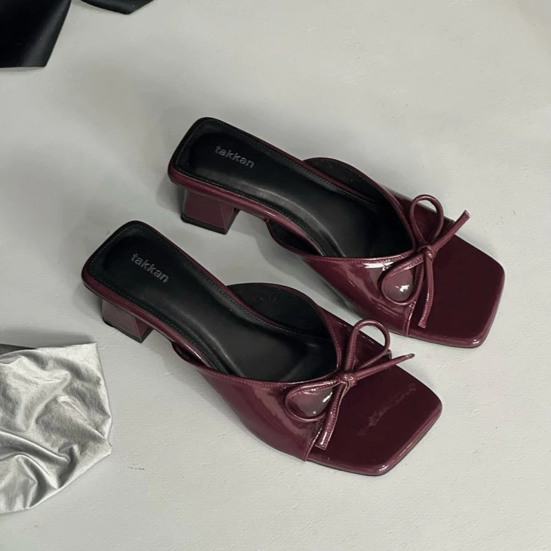 Pair of maroon high-heeled sandal with bow details by Takkan on a light gray background