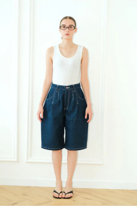 Person wearing a white tank top and blue denim barrel short pants standing against a white wall.