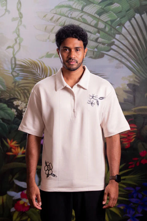 Man wearing a white polo shirt with floral embroidery designs by Nona against a tropical background