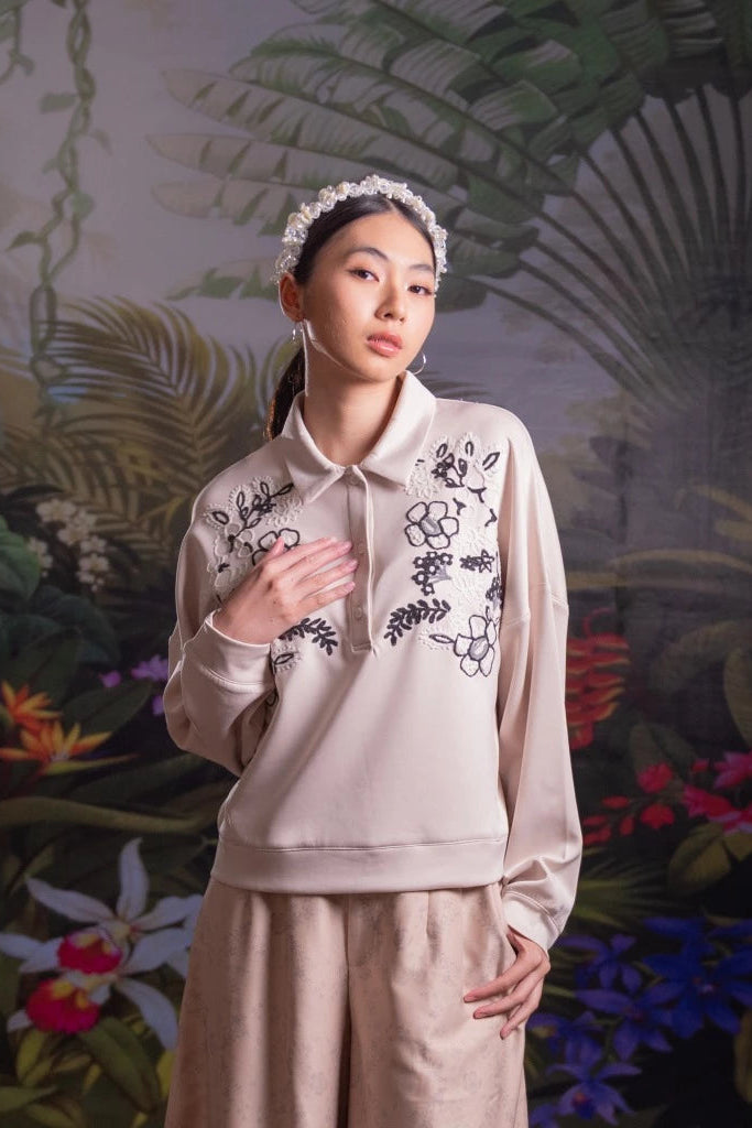 Woman wearing a beige longsleeve polo shirt with floral embroidery by Nona against a tropical background