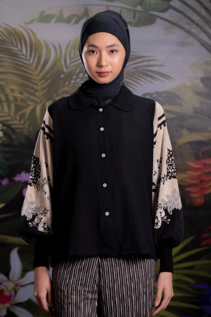 Person wearing a black sweater with patterned sleeves by Nona against a tropical plant background