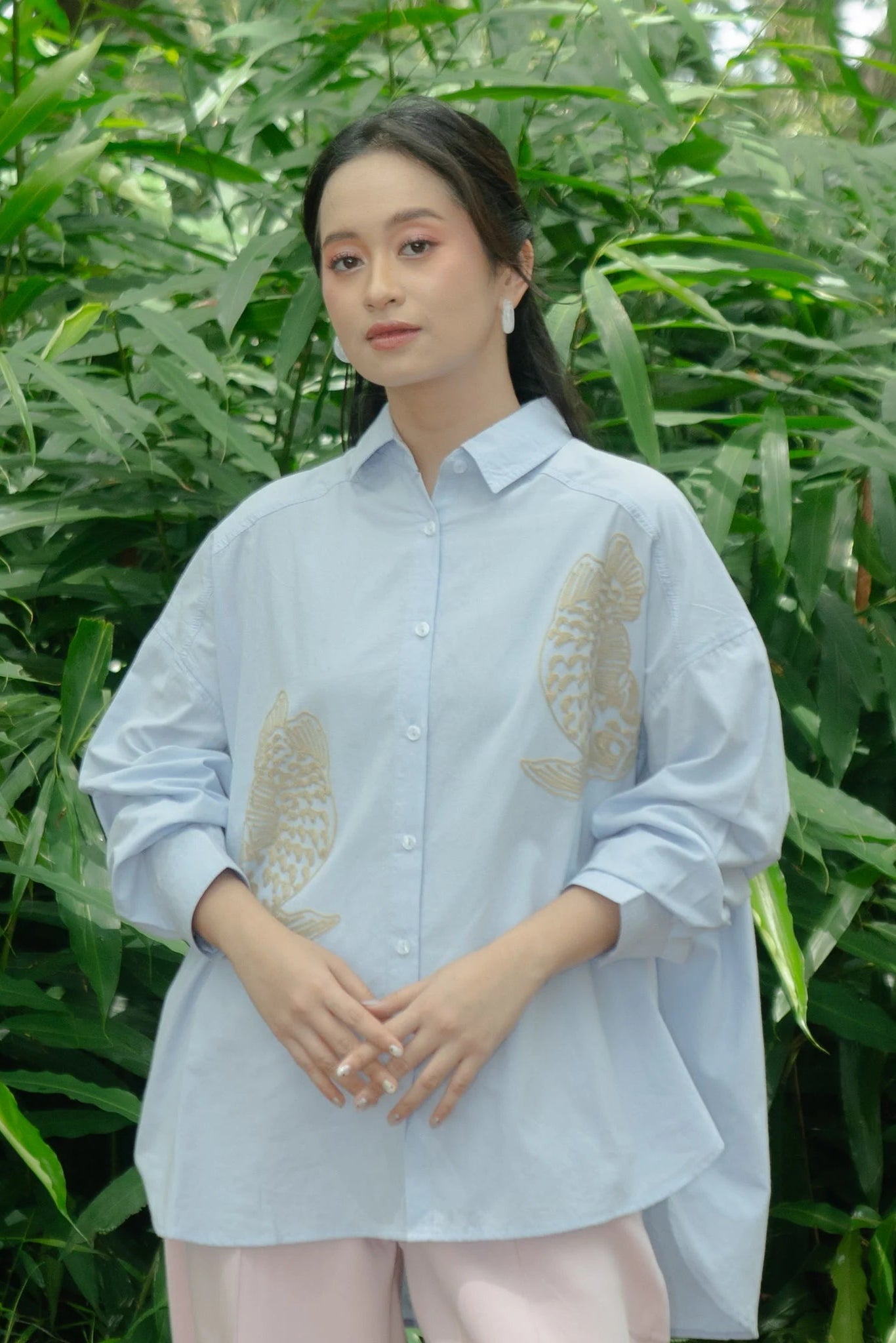 Woman wearing a light blue shirt with gold embroidered fish design by Nona in front of green foliage