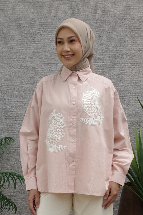 Woman wearing a light pink shirt with white fish embroidery by Nona against a gray wall.