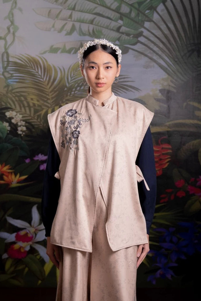 Woman in a beige vest with floral embroidery by Nona standing against a tropical plant background