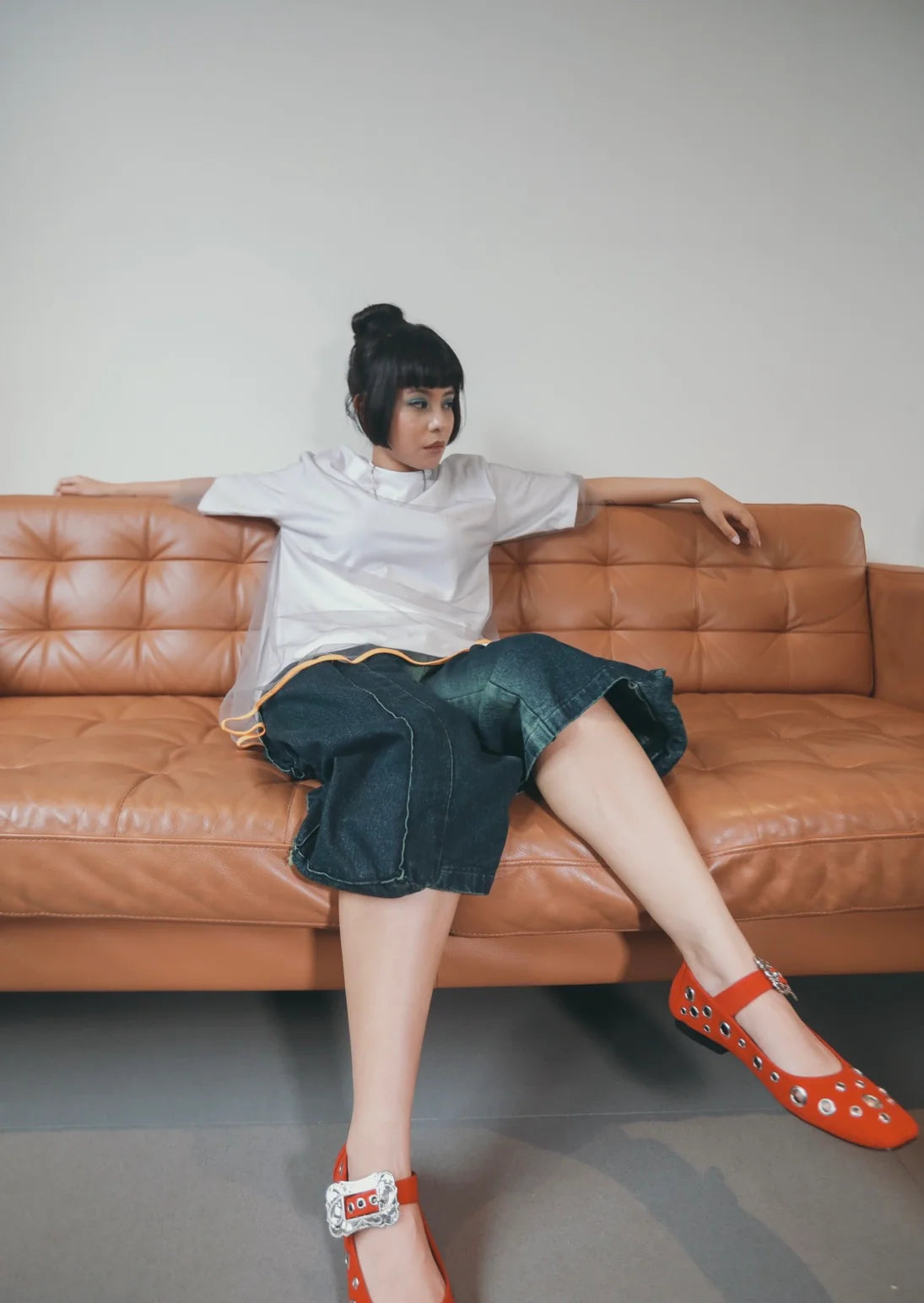 A person seated on a brown couch wearing a white shirt and denim shorts by Wad Studio with a painted blue wash, paired with red floral sandals.