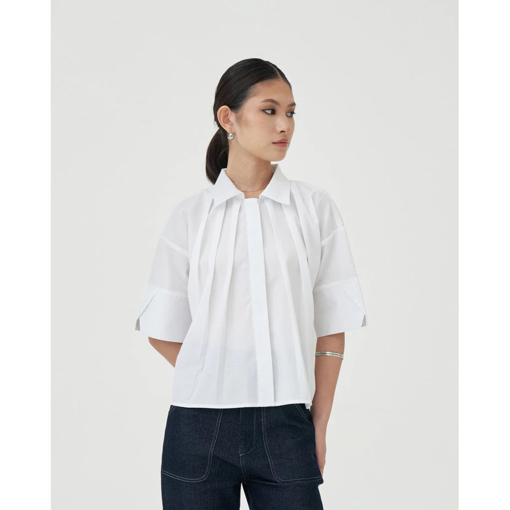 A woman wearing a white blouse with short sleeves and a round neckline by Achete de Nous, paired with white pants.