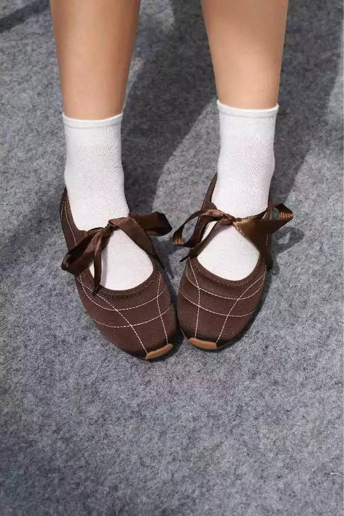 Brown flatshoes with ribbons worn with white socks on a gray floor.
Flat shoes wanita berwarna coklat yang dikenakan dengan kaus kaki putih di atas lantai abu-abu