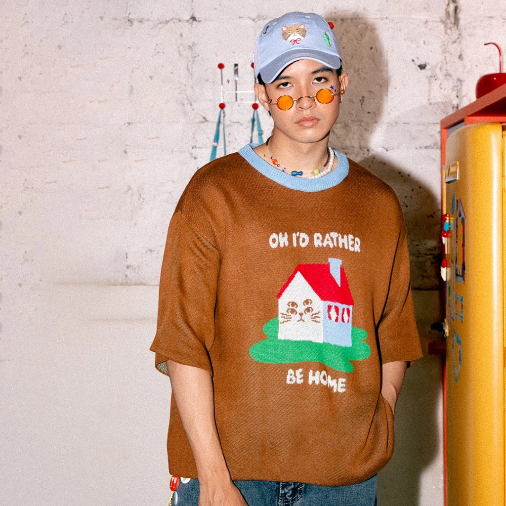 Person wearing a brown t-shirt by Oh.Irv with a house graphic and text, standing against a white brick wall.