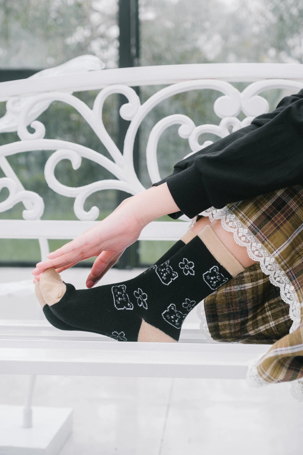 Black crew socks by Tome Ame with white cartoon bear and clover patterns, worn on a person's feet while sitting on a white bench.