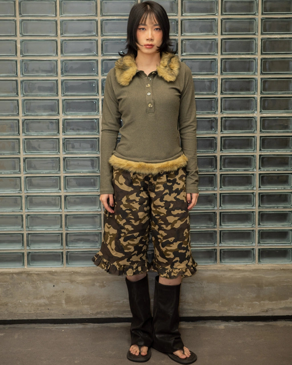 Person wearing a green top with fur trim and camouflage jorts pants against a glass block wall.