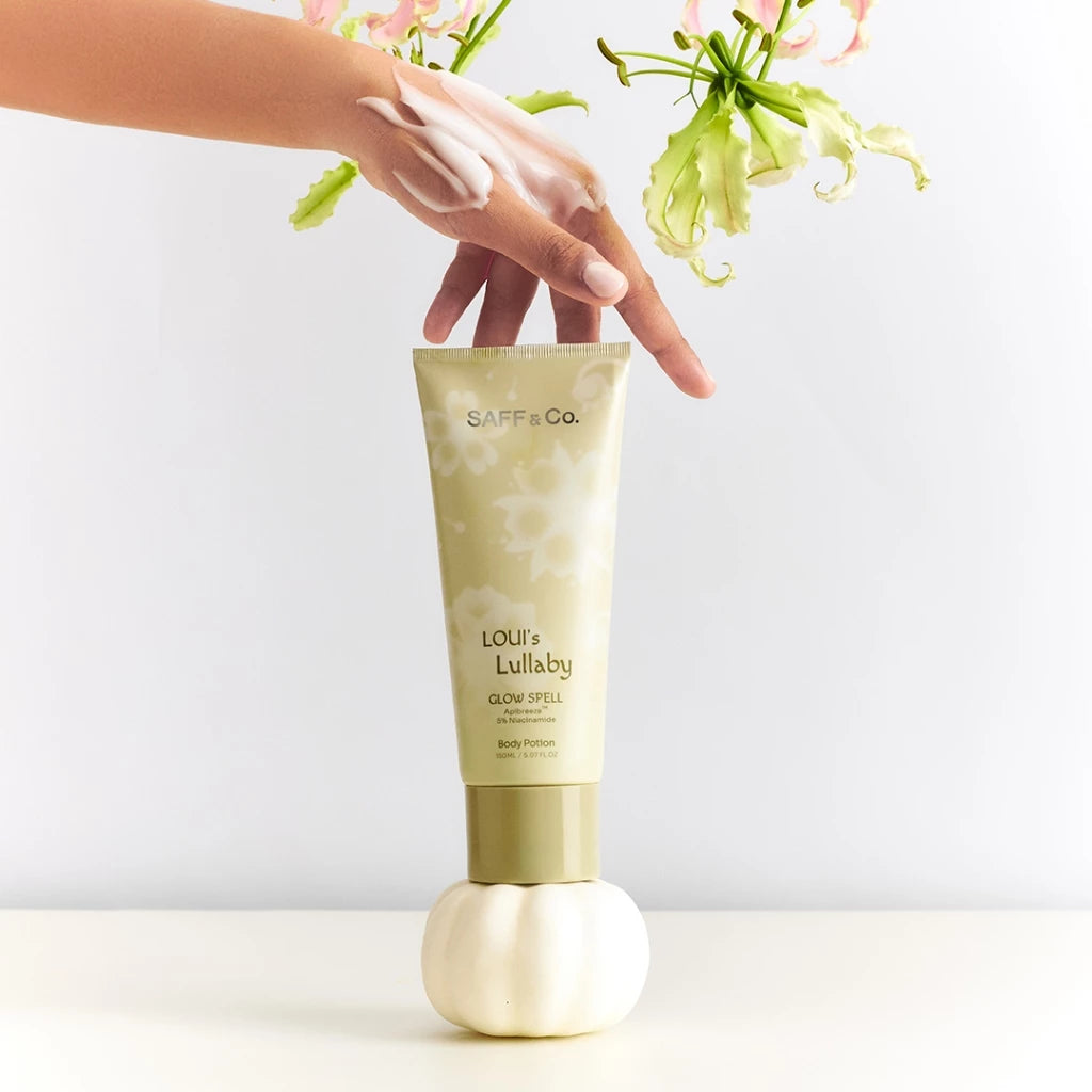 A person's hand holding a pump bottle of Saff & Co. Body Potion - Loui's Lullaby with a floral background.