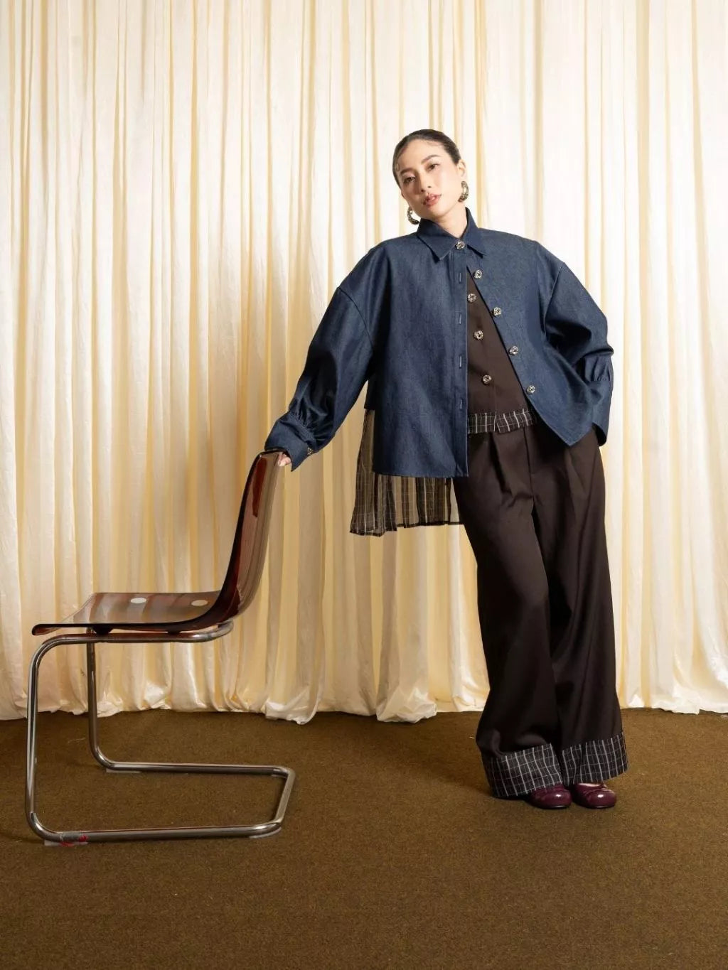 Person wearing a blue denim jacket by LR Studio and dark pants standing next to a chair against a beige curtain background
