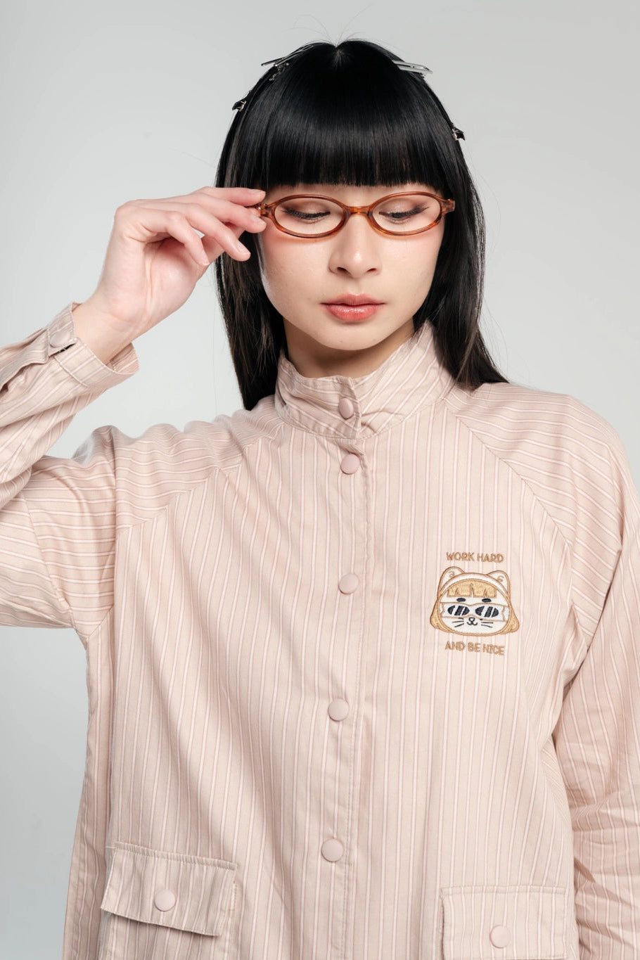 A model with black hair and bangs wearing a light beige pinstriped jacket by Tome Ame with a bear graphic on the chest, holding a pair of tortoiseshell glasses against a light gray background.