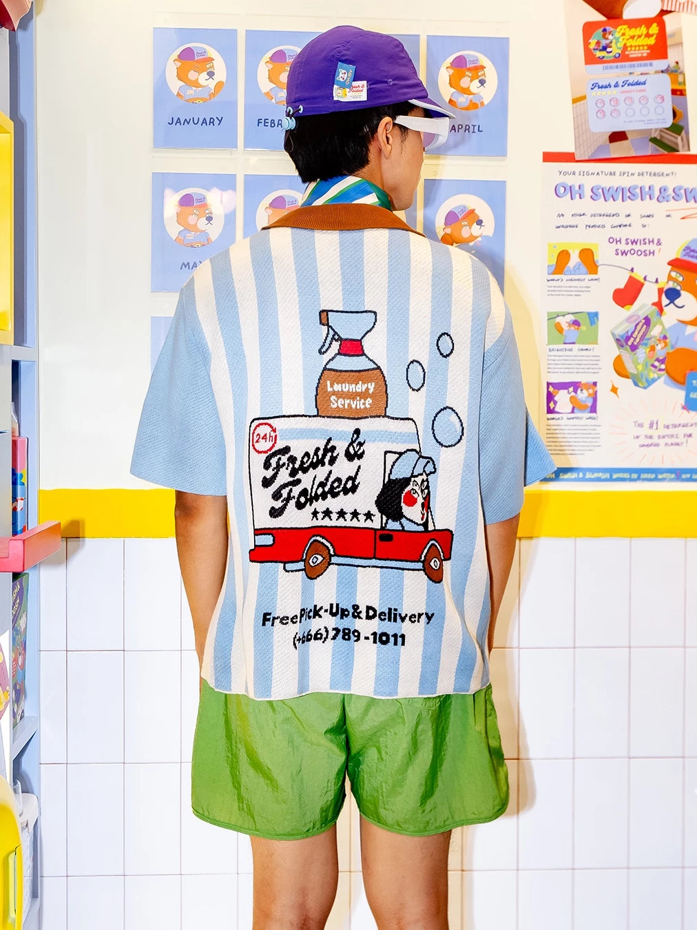 Person wearing a striped knitted shirt by Oh.Irv with a cartoon design at the back and green shorts in front of a colorful wall.