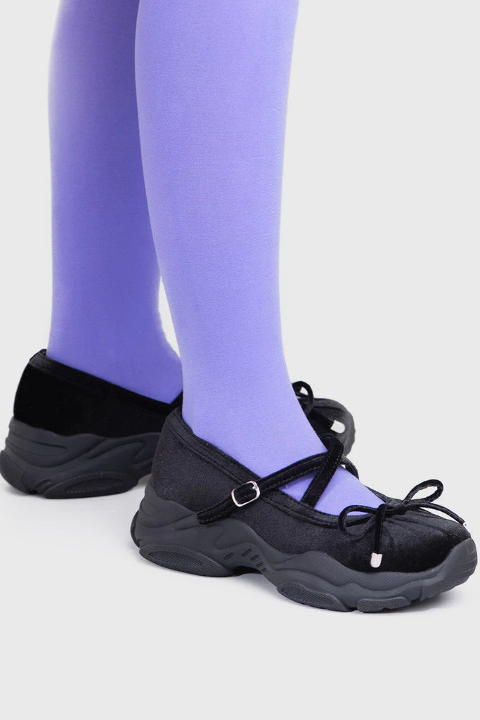 Black ballerina shoes by Adorable Projects with a bow worn with purple tights on a light gray background