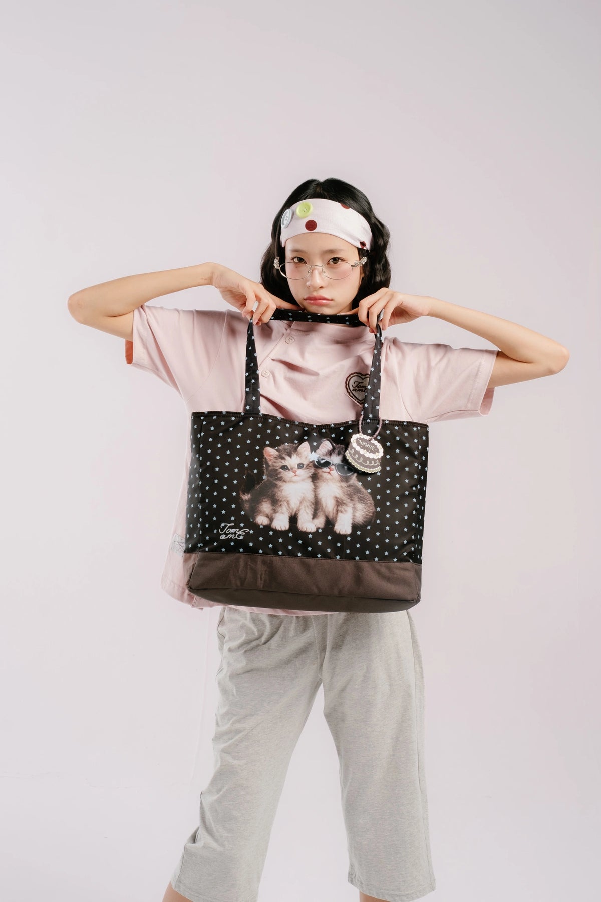 A model wearing a pink t-shirt and gray sweatpants holds up a brown tote bag by Tome Ame with white polka dots and a printed image of two kittens on the front, set against a plain light gray background.