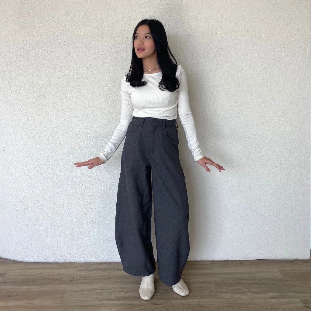 Woman wearing a white top and grey barrel pants standing against a plain wall.
Seorang wanita yang mengenakan atasan putih dan celana barrel berwarna abu-abu sedang berdiri di depan dinding polos.