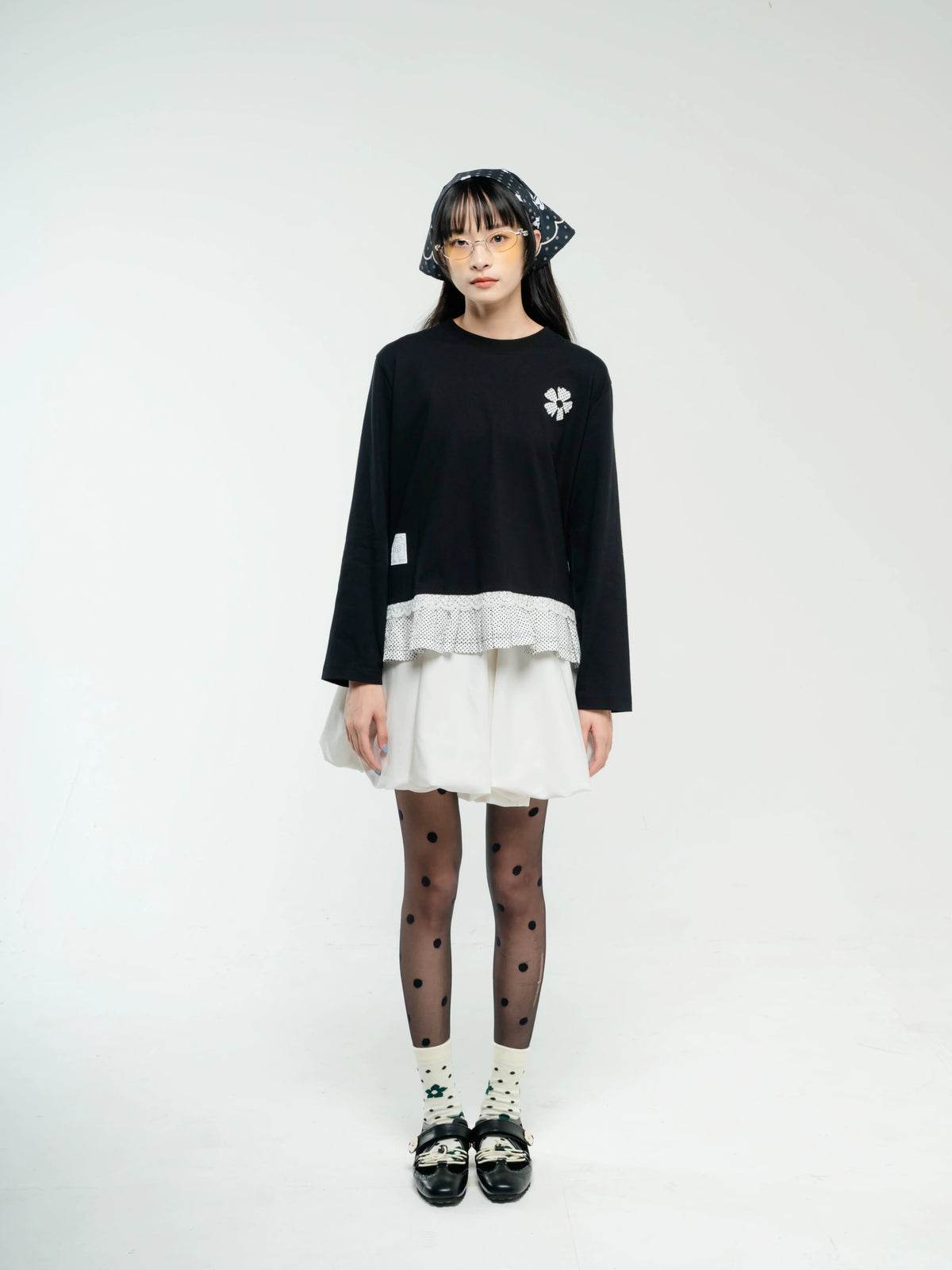 A young girl stands against a plain white studio background wearing a black long-sleeve top by Tome Ame with a white ruffled hem, a white skirt, black polka-dot tights, white socks, and black shoes. She also wears a dark patterned headband.