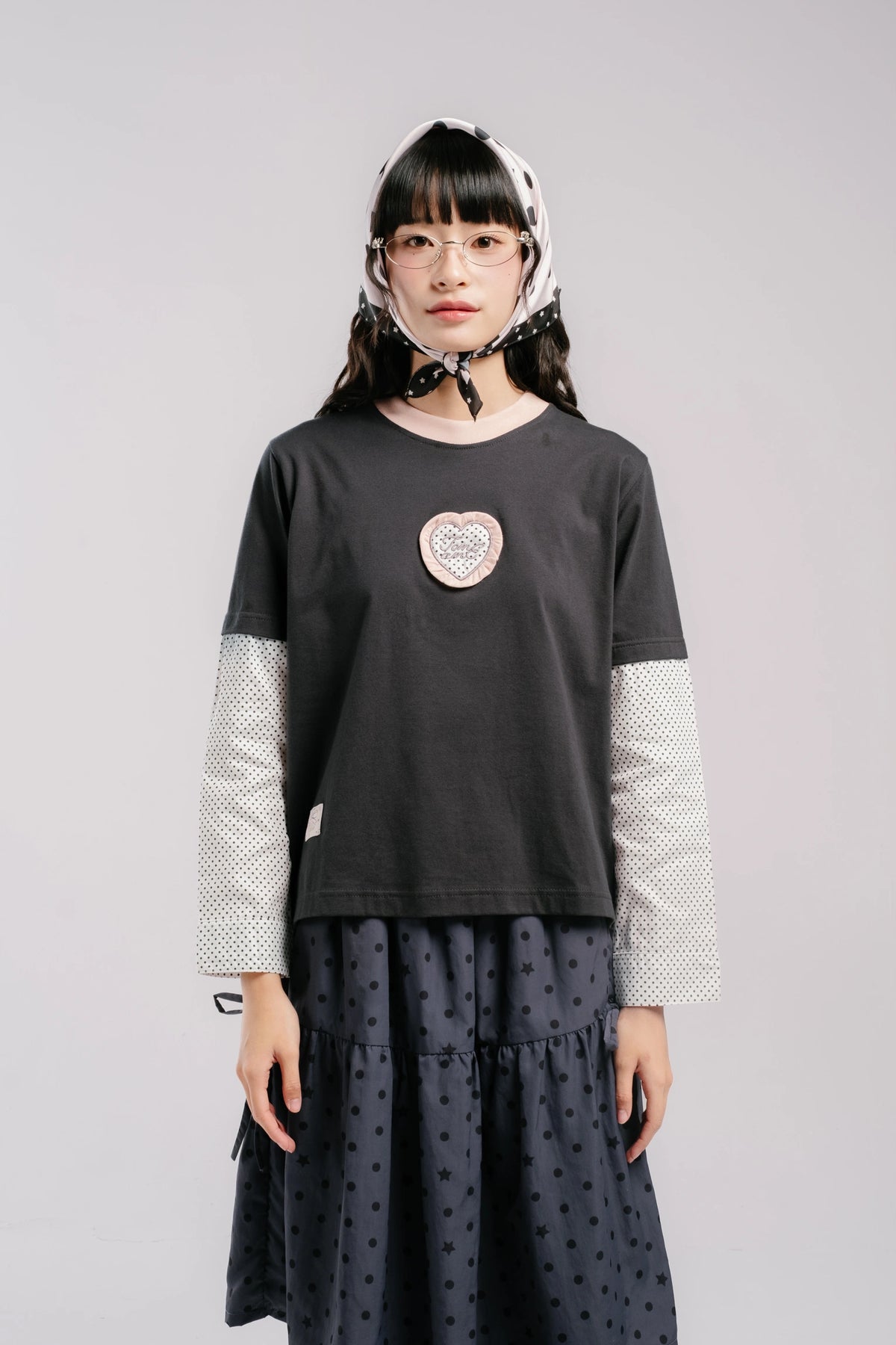 A model wearing a black long-sleeve top by Tome Ame with white patterned sleeves, a heart-shaped patch on the chest, and a dark polka-dot skirt, set against a plain light gray studio background.