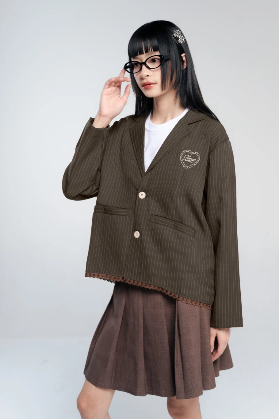 A model wearing a dark brown pinstriped blazer by Tome Ame with a heart-shaped embroidered patch on the chest, paired with a matching pleated skirt and a white t-shirt.
