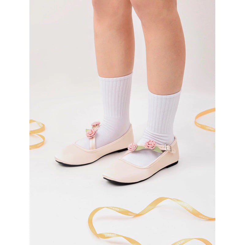 A woman wears cream flatshoes with pink rose embellishments and ankle strap, paired with white ribbed socks, perfect for formal and casual occasions.