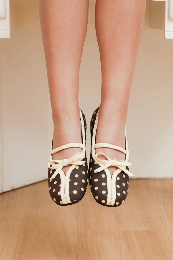 A close-up of a person's legs hanging in the air, wearing brown and white polka dot ballet flatshoes by Mar & Ma with cream-colored straps, set against a warm beige background with white furniture and a wooden floor.