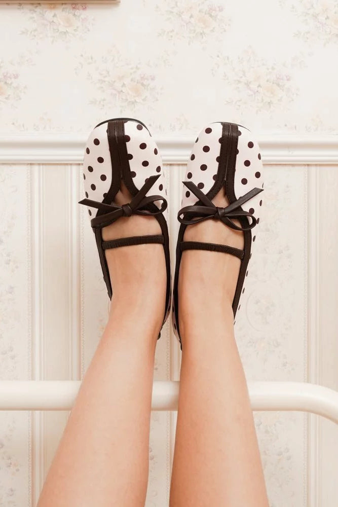 A person's legs are stretched up to a white bed frame wearing pink and brown polka dot ballet flatshoes by Mar & Ma with brown ribbon bows.