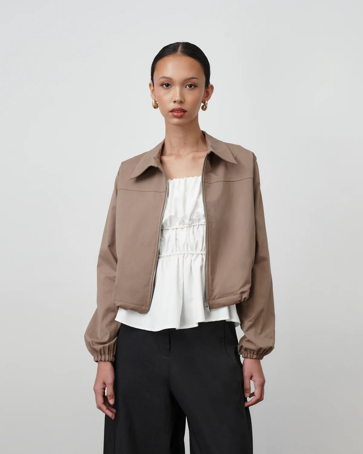 Woman wearing a brown Anders Jacket by Acheté de Nous over a white top and black pants on a light gray background