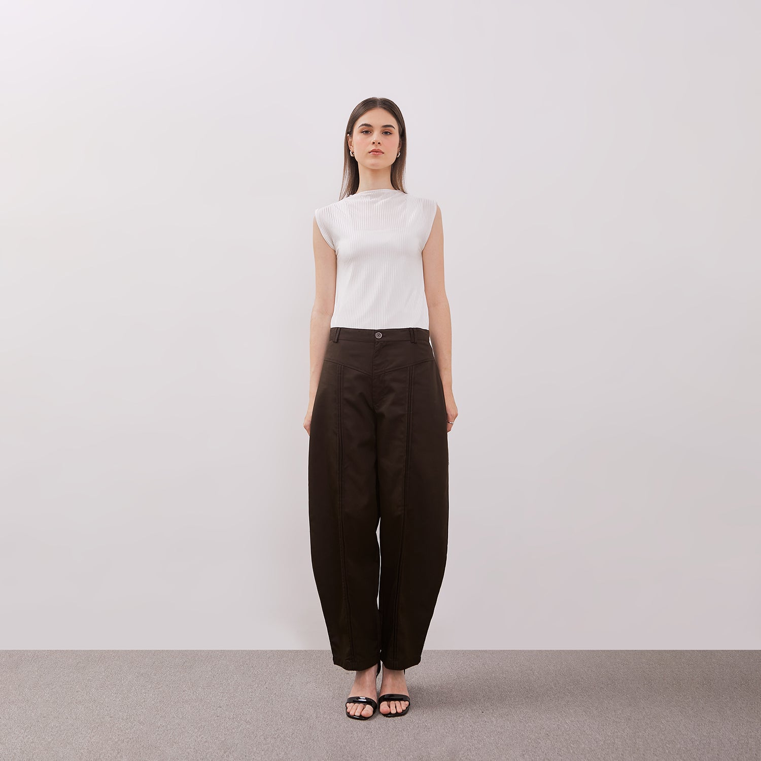 Woman wearing a white sleeveless top and dark brown barrel pants against a plain background