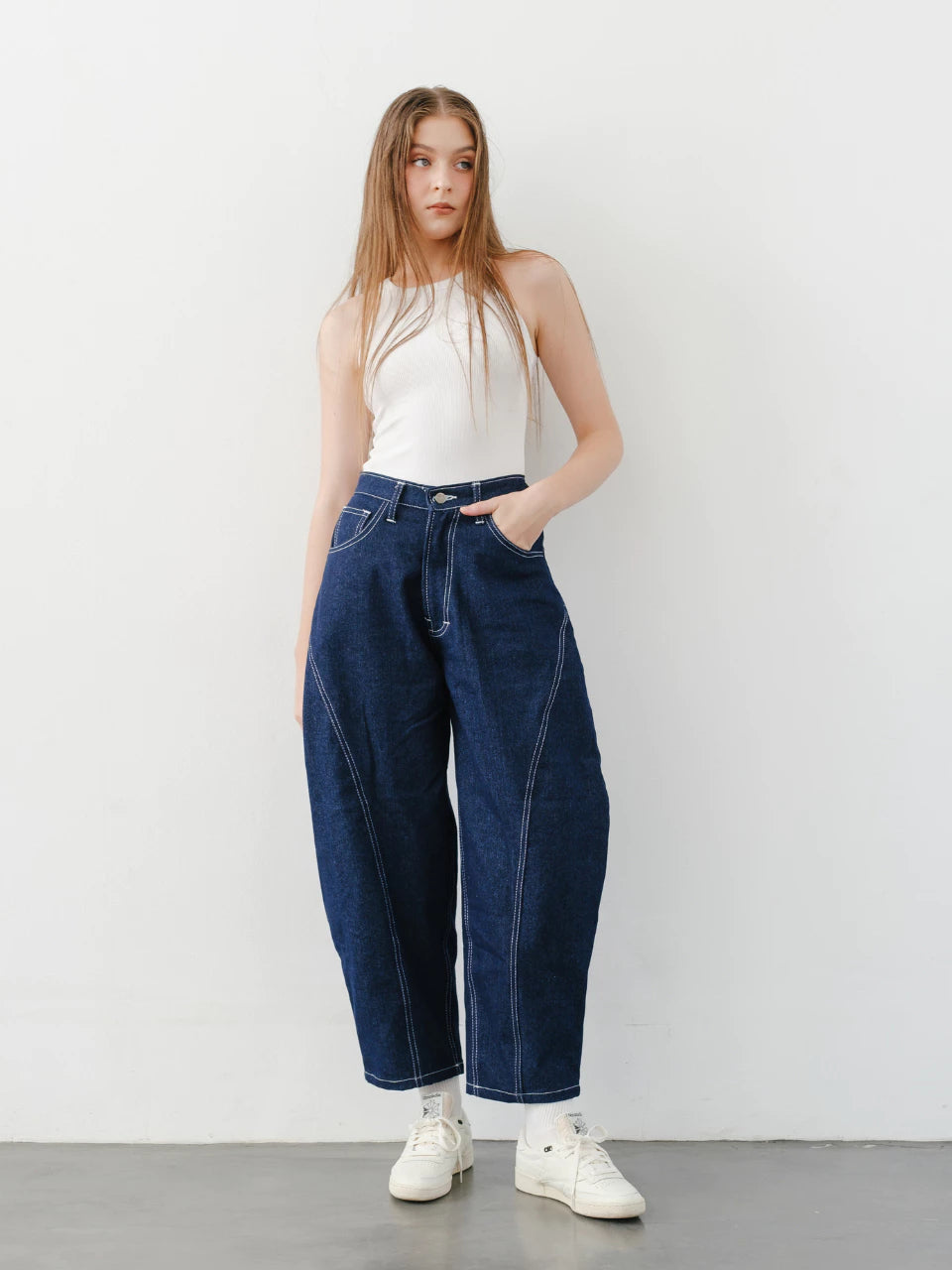 A female model standing against a white background, wearing a white tank top and blue denim barrel pants with a relaxed fit.