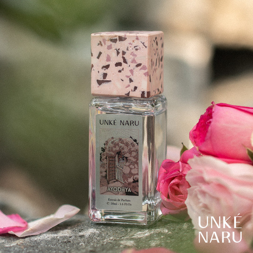 A bottle of 'Ayodhya' perfume by Unke Naru, placed amidst pink roses with a blurred background.