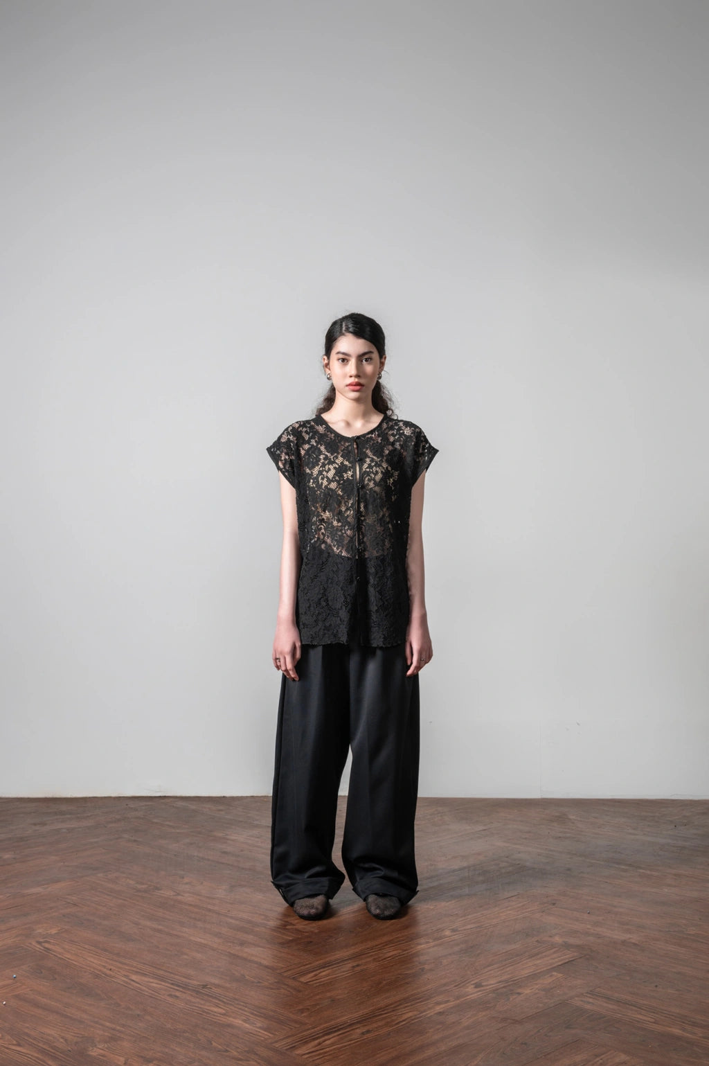 Person wearing a black lace vest made by embroidery floral with a sculptural silhouette and black pants on a plain background