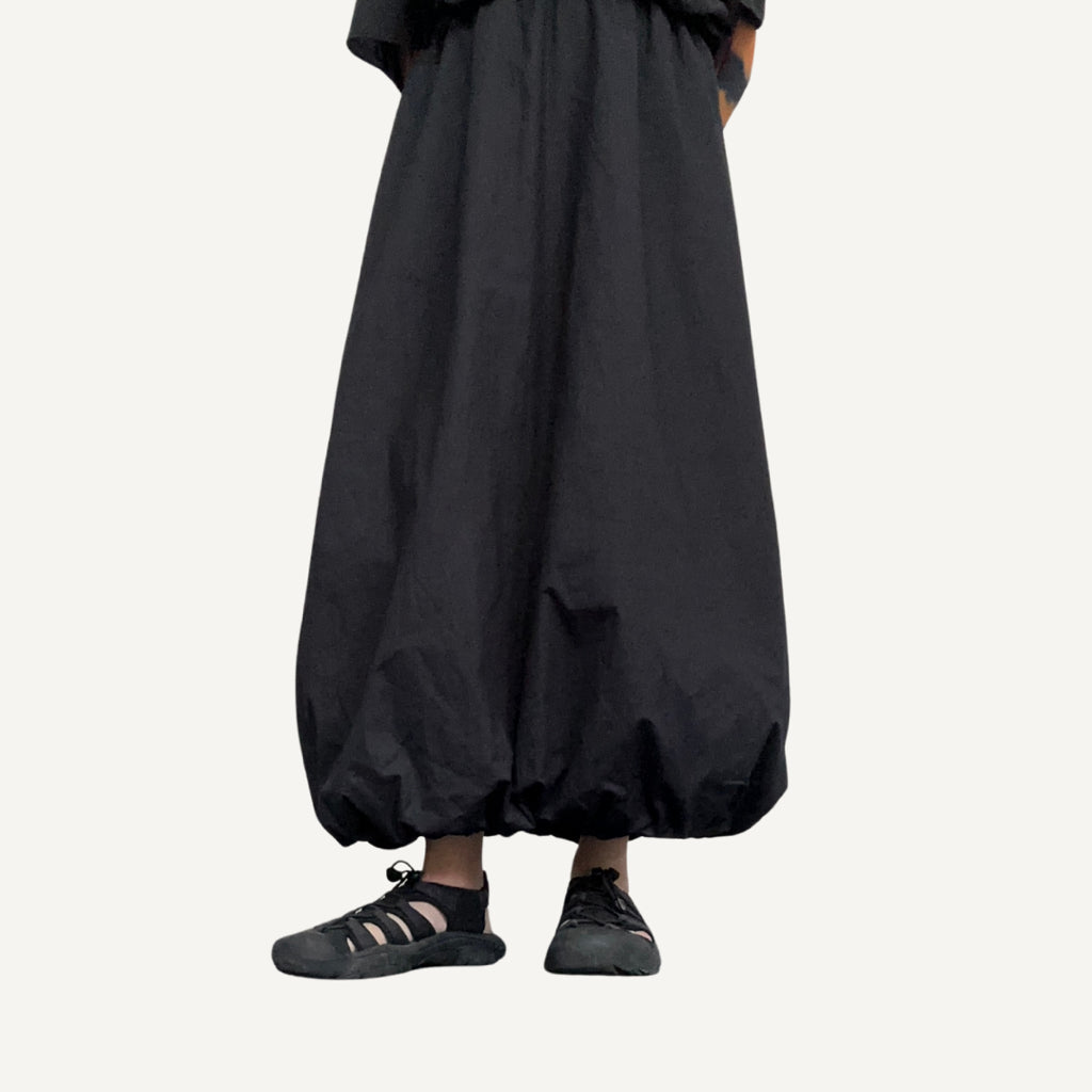 A person standing wearing a black balloon puff skirt with a full rubber waistband and a pair of black sandals.