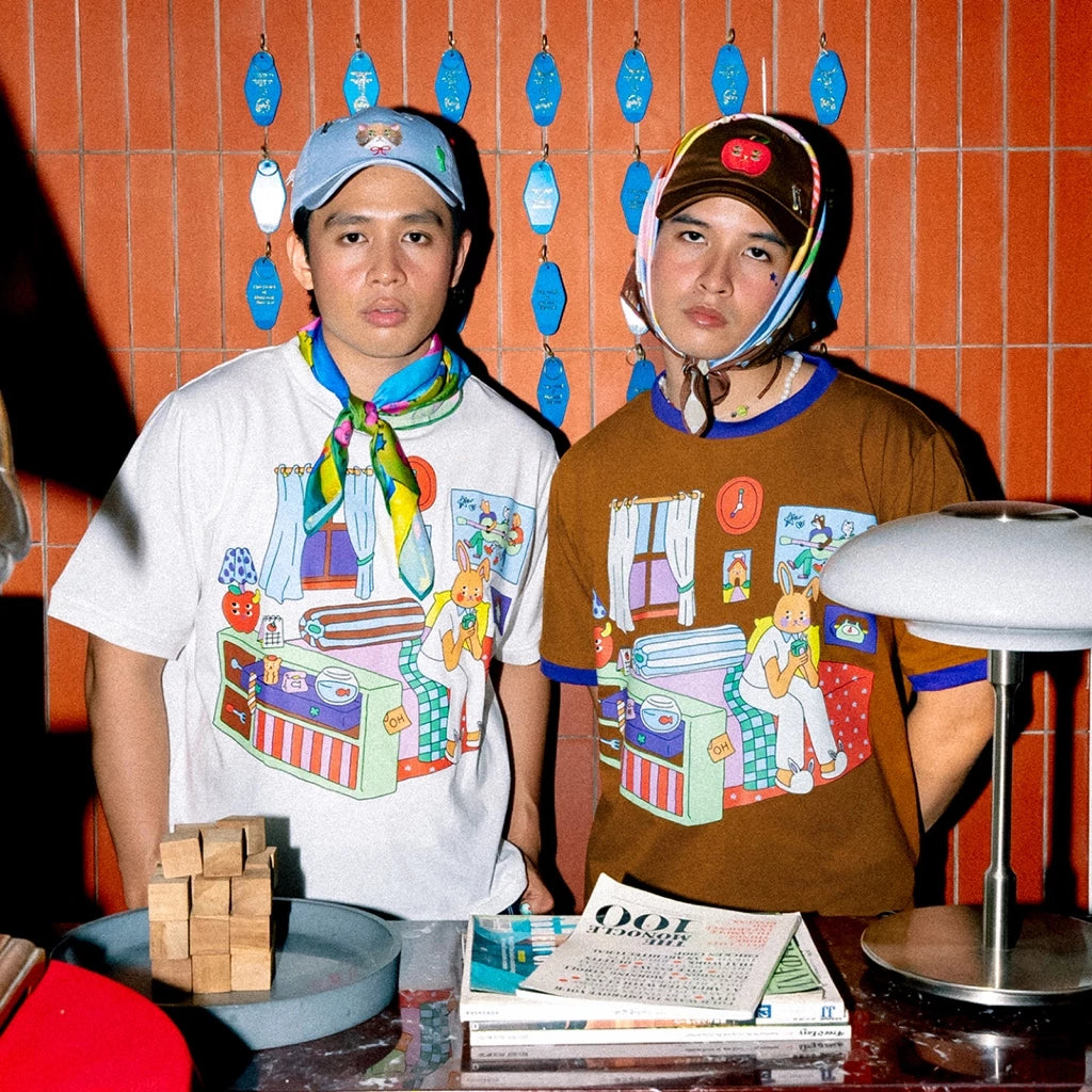 Two people wearing colorful t-shirts by Oh.Irv with cartoon designs in a room with orange walls and decorative items.