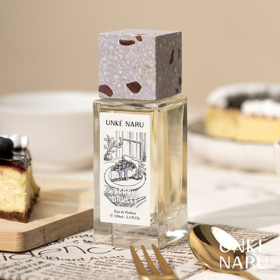 A bottle of Berry Brulee perfume by Unke Naru, presented on a surface with a piece of cheesecake and a fork, against a soft-focus background.