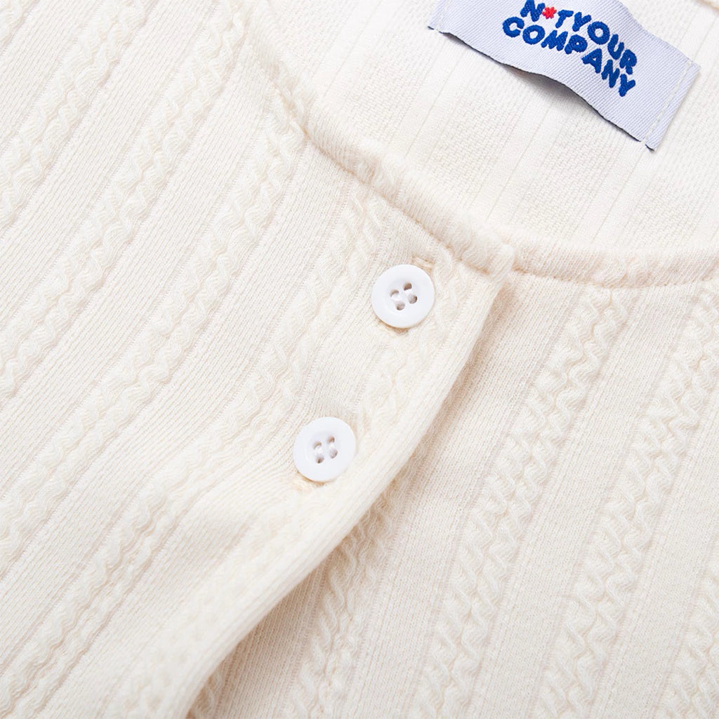 Close-up of a cream-colored textured cardigan with buttons and a label by notyourcompany on a white background