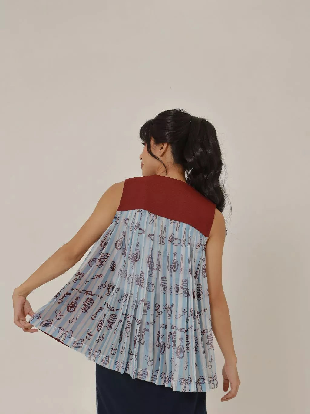 Back view of a woman wearing the Bora Vest Sheer Adult in Wine by LR Studio. The vest features a sleeveless design with a maroon upper panel and a flowy, pleated sheer fabric in light blue with whimsical printed motifs.