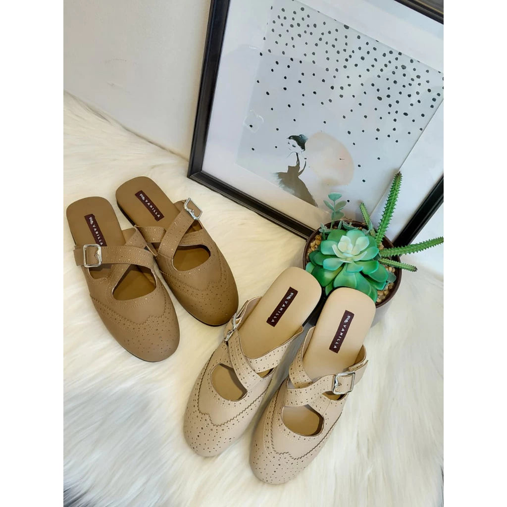 Pairs of cream and brown mules on a light wooden floor with a decorative mirror and plant in the background.