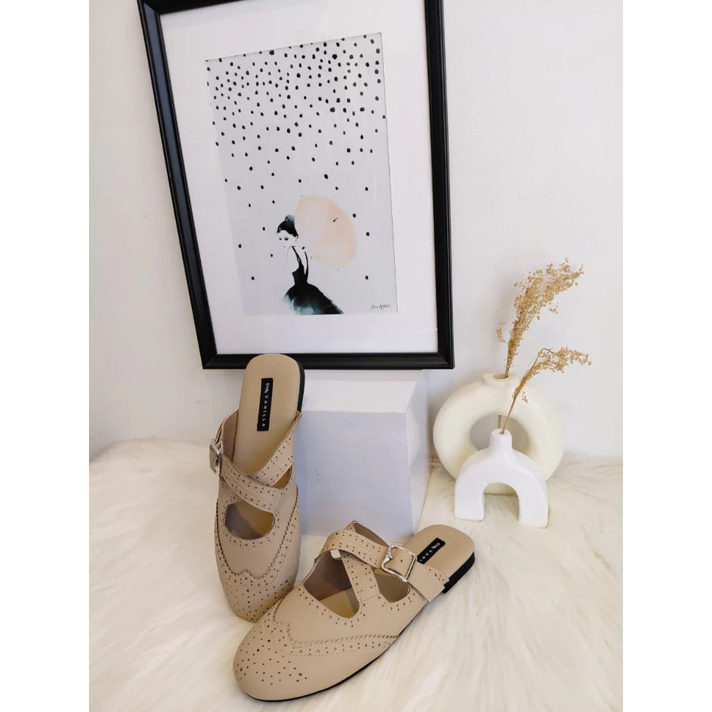 Pair of brown mules on a white surface with a framed artwork in the background.