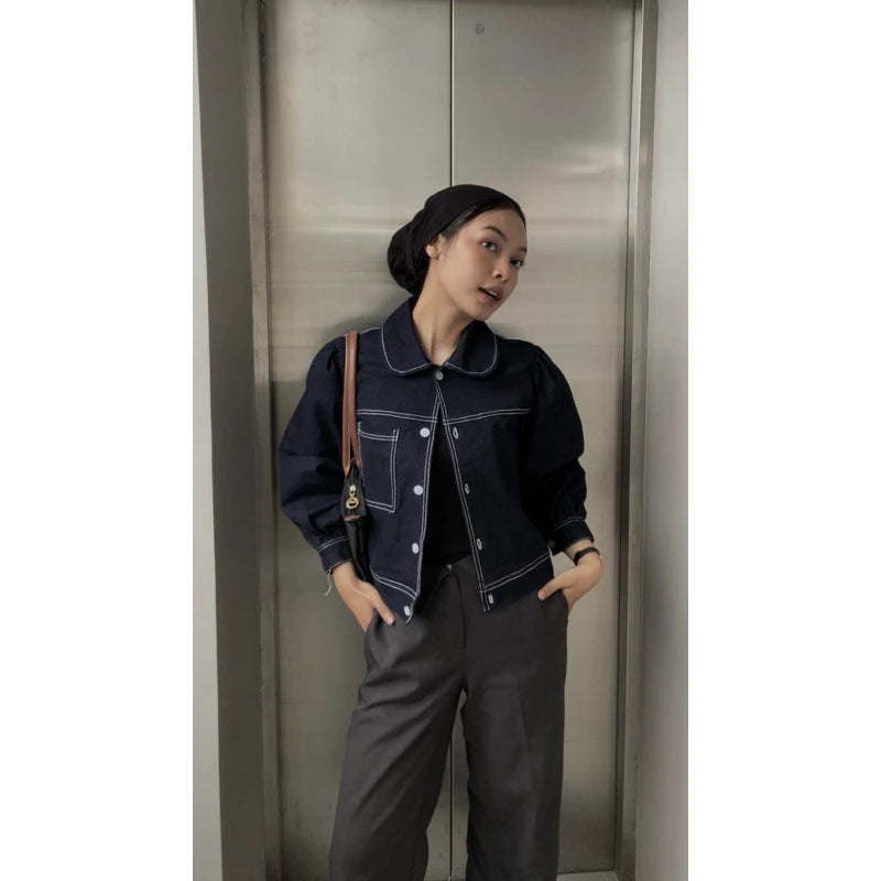 Person wearing a dark denim jacket by Not Your Company and gray pants standing against a metallic elevator wall.