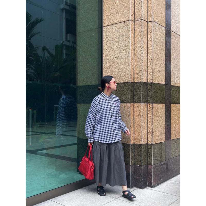 Woman in checkered Brixton shirt by notyourcompany and gray skirt holding a red bag against a modern building.